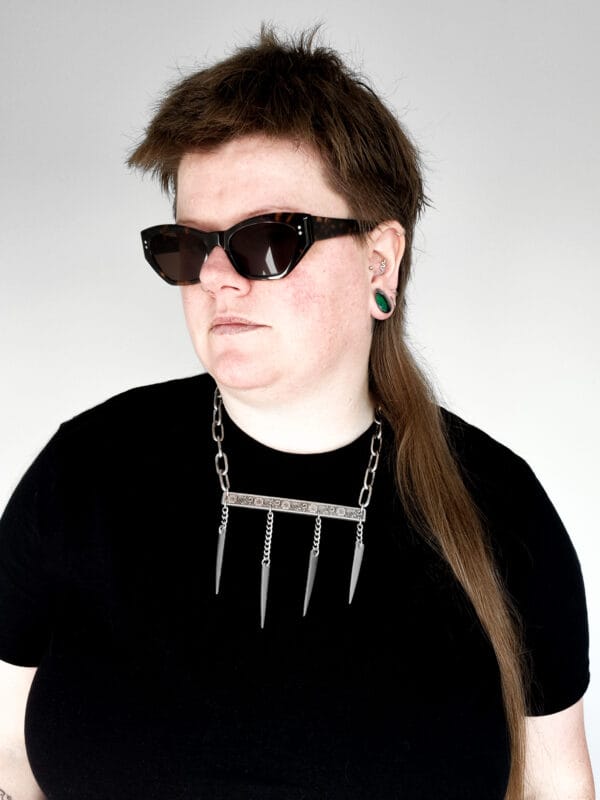 a woman wearing sunglasses, a black shirt and a stainless steel necklace made from the handle and fork tines of a vintage fork.