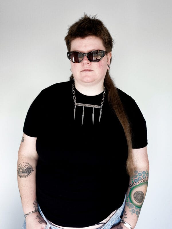 a woman wearing sunglasses, a black shirt and a stainless steel necklace made from the handle and fork tines of a vintage fork.