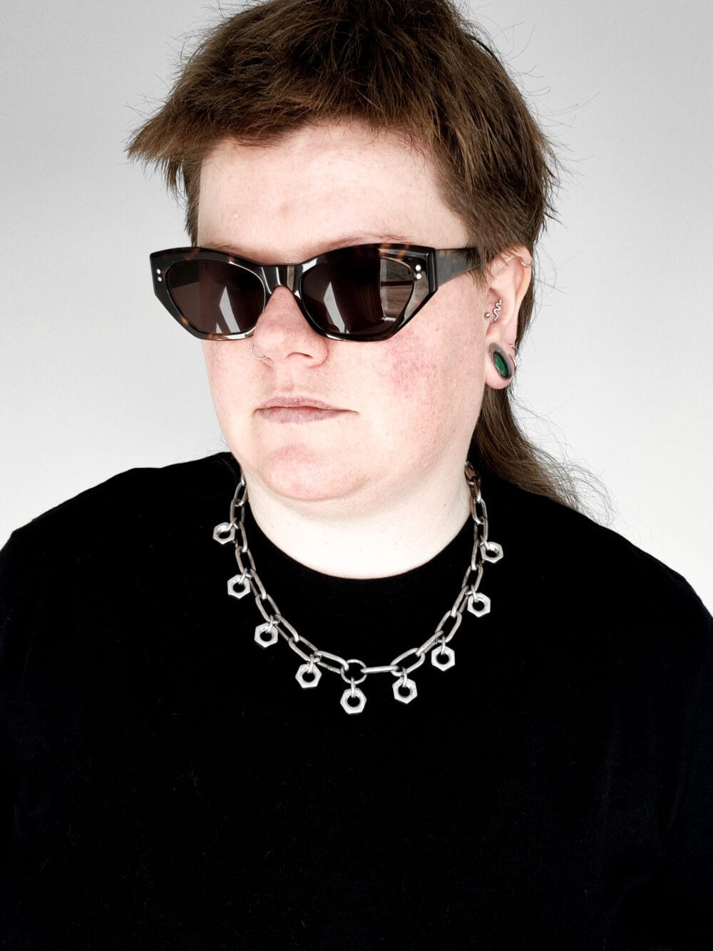 a woman with sunglasses and a black shirt wearing a necklace made from chunky stainless steel chain and hex nuts as charms along it.