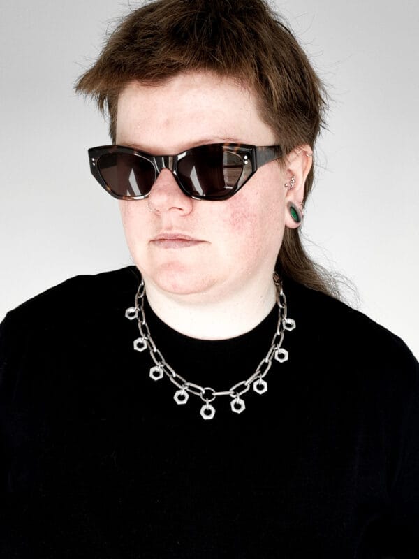 a woman with sunglasses and a black shirt wearing a necklace made from chunky stainless steel chain and hex nuts as charms along it.
