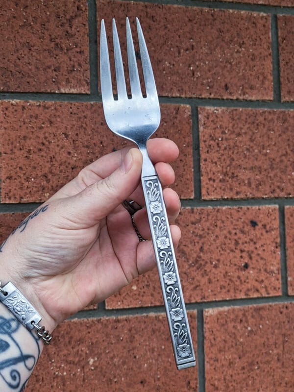 an image of a hand holding a giant vintage fork. the handles have a flower and swirly leaf pattern