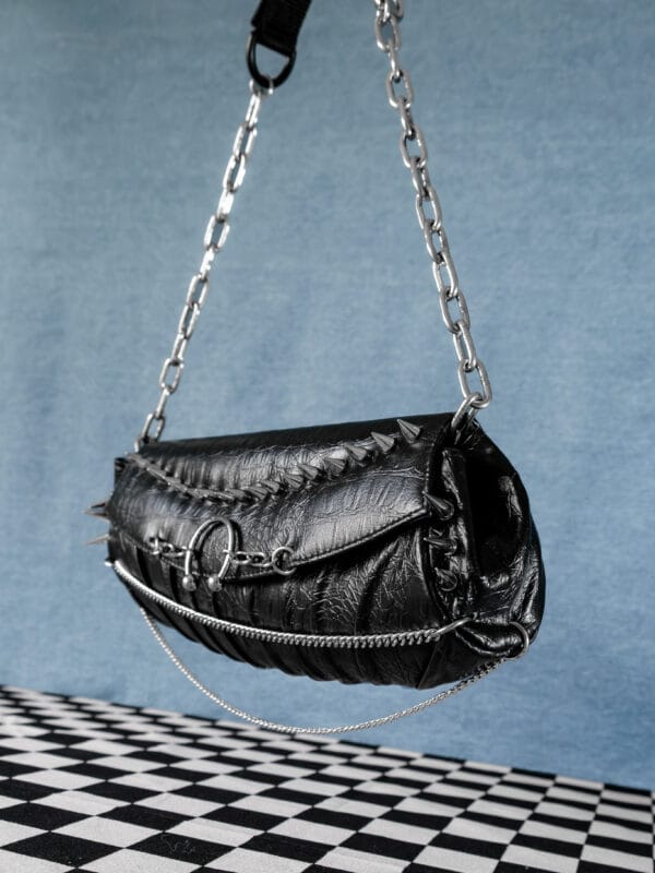 a product photo of a black faux leather handbag with spikes and chains.