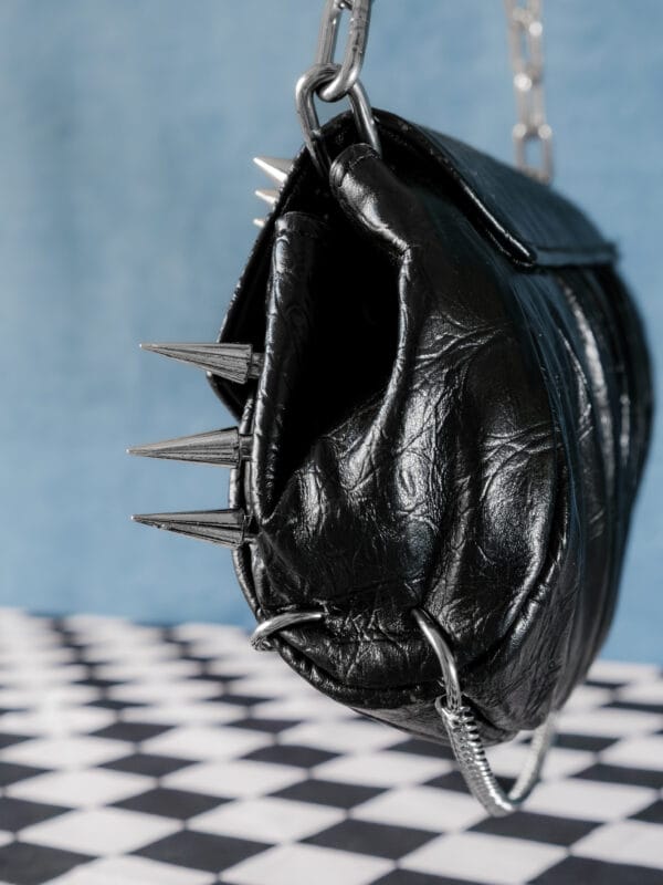 a product photo of a black faux leather handbag with spikes and chains.