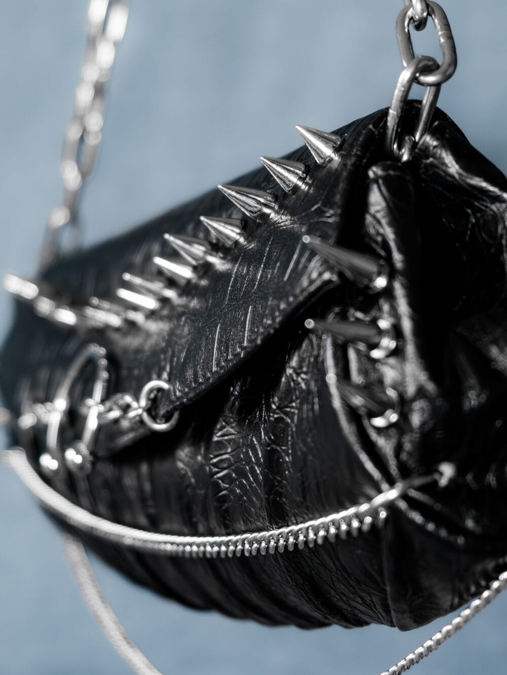 a product photo of a black faux leather handbag with spikes and chains.