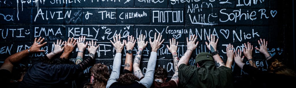 a group photo from a brand photoshoot where 7 people are reaching up and placing their hands on a brick graffiti wall to show the rings on their fingers.